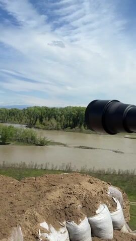 The video presents a panoramic view of a wide, muddy river landscape under a bright, partly cloudy sky. The camera pans slowly from left to right, revealing different elements of the scene.

In the foreground, there's a steep, eroded dirt bank, with several white sandbags partially visible, possibly used for reinforcement or as a makeshift barrier. Below this bank, the river flows, appearing wide and somewhat turbid, with visible sandbars or small islands covered in sparse vegetation. Lush green trees line both sides of the river, forming dense forests that stretch into the mid-ground.

As the camera pans further right, distant industrial structures come into view on the horizon, including tall chimneys and cooling towers, suggesting the presence of a power plant or similar facility. Beyond these structures, a range of mountains, possibly snow-capped, can be seen in the far distance, blending with the clouds. The sky is a vibrant blue with scattered white, wispy clouds. The overall impression is one of a natural river environment contrasting with distant industrial development. A dark object, possibly part of the recording device, is visible in the right foreground at the beginning of the video.