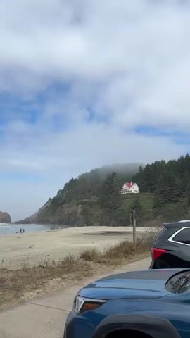 The video shows a scenic view of a beach with a wooded hillside in the background. A white house with a red roof is visible on top of the hill, partially obscured by fog. There are several cars parked in the foreground. The beach is sandy, and there are a few people walking along the shore. The ocean is calm, and the sky is partly cloudy with patches of blue. The overall atmosphere is peaceful and serene.