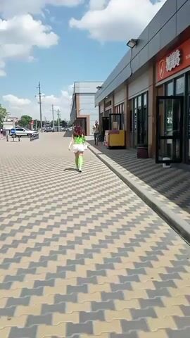A woman with green hair wearing a green top and white shorts walks away from the camera down a checkered paved walkway. She is walking towards a building with large glass windows and doors. A man is standing outside the building. The sky is blue with scattered white clouds.