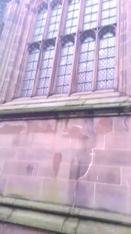 The video shows a low-angle view of the exterior of a large, historical building with a prominent arched window. The window is divided into multiple smaller panes in a diamond pattern. The building is constructed of reddish-brown stone, and there are decorative carvings and statues on the facade. The sky is overcast, and the upper portion of the building appears to fade into the mist.