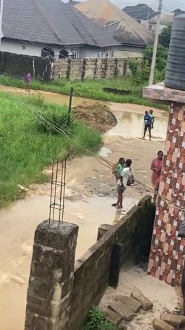 The video shows a flooded street with several people. In the foreground, a person is standing near a wall holding a child. To their right, another person in a pink garment stands and looks towards the left. In the middle ground, a person is walking across the flooded street with a child on their back. Further back, two children are walking together through the water. In the background, a person in a purple garment is walking away from the camera on a drier patch of land. The environment appears to be a residential area with houses and overgrown vegetation. The sky is overcast. The water is muddy and appears to be several inches deep in some areas. Overhead wires are visible, supported by poles. A concrete pillar with exposed rebar is in the lower left of the frame. A water tank is visible on the right side of the frame.