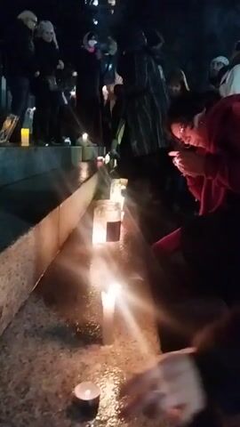 A group of people stand on a set of stone steps, some holding candles, at night. The steps are lined with lit candles, some in glass jars, and some on the stone surface. A woman in a red jacket lights a candle with a lighter. Another person stands next to her, holding a white candle. The area is lit by the candles and streetlights. The mood appears somber.