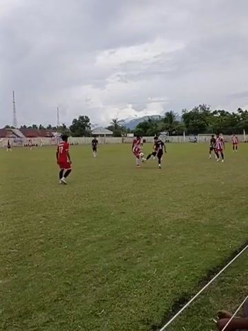 A soccer game is being played on a grassy field. There are several players on the field, some in red and white uniforms and others in black and white uniforms. The sky is cloudy. In the background, there are buildings and trees. One player in a red uniform with the number 17 is in the foreground, dribbling the ball. Another player in a black uniform is attempting to tackle him. Other players are scattered across the field, some watching the action, others moving into position. The game appears to be in progress, with the ball in play.