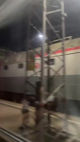This media file captures a nighttime scene at a train station in Inza, Russia. The primary subject is a red and gray electric locomotive, identified by the number "H929486" visible on its side. The train is stationary at the platform, viewed through a window, suggesting the footage was taken from another train or a waiting area.

The setting is an outdoor train station platform, illuminated by artificial lighting that casts a dim glow on the tracks and surrounding infrastructure. A metal support structure, likely part of the station's overhead canopy or electrical system, dominates the right side of the frame. A yellow warning sign with a black symbol of a train and an exclamation mark is affixed to this structure, and beneath it, text in Russian reads "Берегись поезда!" which translates to "Beware of the train!".

Above the train, a faint white plume of smoke or steam is visible against the dark night sky, adding a somewhat atmospheric element to the scene. The overall mood is quiet and perhaps a bit melancholic, characteristic of a late-night railway station. There are no people clearly visible in the frame, but shadows on the platform and near the support structure suggest their potential presence. The reflection on the window creates a slightly hazy effect, softening the details of the image. The time of day is definitively night.