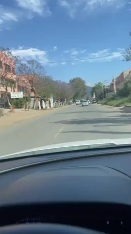 The view from inside a car shows a two-lane road with moderate traffic. Trees line the left side of the road, some with purple blossoms. Buildings and shops are visible behind the trees. A white van with blue items on its roof drives towards the camera, followed by a black SUV. The sky is blue with scattered clouds. Power lines run along the right side of the road. The road is slightly uneven with visible patches and lines. A mountain range is faintly visible in the distance. The car's dashboard is in the foreground.