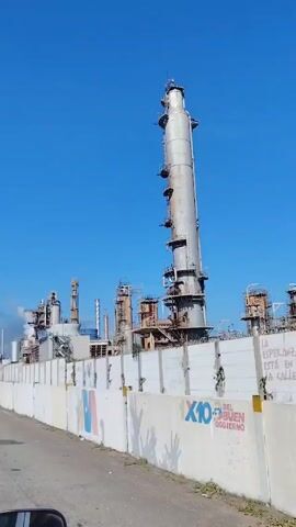 The video shows a large industrial complex with a tall, cylindrical tower and several other structures, likely a refinery or chemical plant. The complex is surrounded by a white concrete wall with graffiti on it. Smoke or steam can be seen rising from some of the structures. The sky is clear and blue. The video is taken from a moving vehicle.