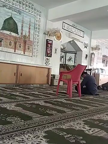 The video depicts the interior of a mosque or prayer hall, likely in Dera Chahal, Pakistan. The room has white walls and ceiling, with a green and white patterned prayer rug covering the floor.

In the foreground, a man wearing a blue shirt and dark pants is seen kneeling on the prayer rug, in the prostration (sujud) position typical of Muslim prayer. A pink plastic chair is placed beside him. Another individual is partially visible kneeling in the background to the right, also engaged in prayer.

The walls are adorned with religious decorations. On the left wall, a large framed tapestry or picture displays a green-domed mosque with two minarets, encircled by Arabic calligraphy. Beneath this artwork is a wooden cabinet with sliding doors. On the wall straight ahead, there is a framed piece of Arabic calligraphy, a digital clock, and another framed item. Two ceiling fans are mounted, and an arched doorway is visible in the background, leading to another area of the building. The scene conveys a sense of peaceful devotion.