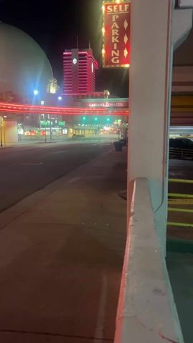 The video captures a nighttime street scene, likely in a city entertainment or casino district, illuminated by numerous neon lights. The camera moves forward slowly, providing a ground-level perspective.

At the beginning (0:00), the view is down a city street. On the right, a prominent "SELF PARKING" sign with red letters, yellow lights, and red arrows pointing downwards is visible, attached to what appears to be a parking garage structure. Across the street on the left, a large, smooth dome-shaped building is illuminated with changing colors. Further down the street, a tall building with vertical red neon lights is visible in the distance. Horizontal red neon lights stretch across the street, and beneath them, a "CAESARS REWARDS" sign with blue and white text is displayed. The street itself is dark asphalt, and a sidewalk is in the immediate foreground.

As the camera pans left and moves forward (0:01-0:03), the dome building becomes more central, and the structures closer to the left side of the street come into view. These include a building featuring wide white and purple vertical stripes, adorned with horizontal red neon lights. Planters with small trees are spaced along the sidewalk. The street remains largely empty of vehicles.

Towards the end of the video (0:03-0:05), the striped building is very close on the left, with more details of its entrance and yellow elements visible. The camera continues moving forward, showing a painted white crosswalk on the street. An entrance to another building, also lit with red neon lights, is visible across the street. The overall ambiance is one of a vibrant, well-lit urban environment that is quiet during the night, with a focus on architectural lighting and signage.