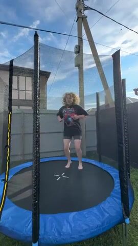 A person with curly blonde hair wearing a dark t-shirt and shorts is jumping on a trampoline. The trampoline has a black jumping surface with a white X in the center and a blue padded edge. The trampoline is enclosed by a black safety net supported by black poles with yellow accents on the left. The background shows a fence, a house, and a utility pole against a cloudy blue sky. The person is captured mid-air, performing a jump with their legs extended.