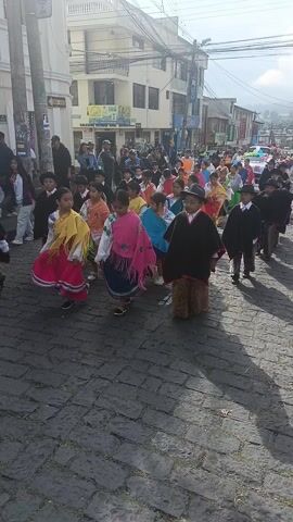A group of children are participating in what seems to be a cultural parade. They are walking down a street paved with cobblestones and the buildings on either side are mostly white with multiple stories. The children are wearing traditional clothing with bright colors and patterns. Some of the boys are wearing black ponchos and hats, and furry leg coverings. Some of the girls are wearing colorful shawls. There are other people watching the parade. The weather looks sunny and bright. Banners are strung across the street.