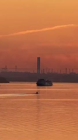 The video begins with a wide shot of a body of water at sunset. The sky is a vibrant orange and red, with a few wispy clouds. A large ferry or cruise ship is visible in the middle distance, moving slowly across the water. In the foreground, a much smaller jet ski is creating a wake as it speeds across the water, leaving a trail behind it. In the background, a long bridge with tall pylons spans the horizon, and a city skyline can be seen beyond it. The water reflects the colors of the sunset, creating a warm and serene atmosphere.