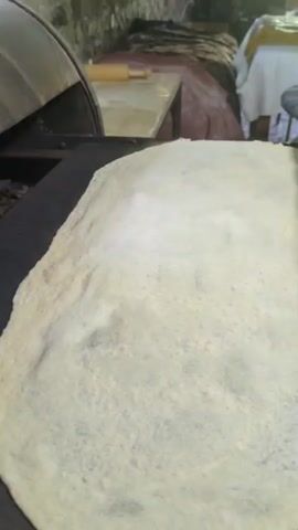The video shows a close-up view of a flatbread being cooked on a hot surface, likely a griddle or tawa. The bread is circular and appears to be made from a light-colored dough, possibly wheat or rice flour. It has a slightly bubbly texture, indicating it is cooking evenly. In the background, a large, curved metal object, possibly a tandoor oven, is visible. A wooden rolling pin rests on a table behind the griddle. A person's hand is seen holding a metal tool with a wooden handle, which is being used to adjust something near the oven. The overall setting appears to be a kitchen or food preparation area, with rough textures and a utilitarian atmosphere. The lighting is somewhat dim, suggesting an indoor environment.