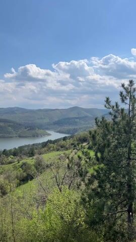The video shows a scenic view of a lake surrounded by green hills under a partly cloudy sky. The camera is positioned on higher ground, offering a wide perspective of the landscape. Trees and bushes are visible in the foreground, partially framing the view. The lake stretches into the distance, winding through the hills, creating a sense of depth in the shot. The sky is blue with scattered white clouds.