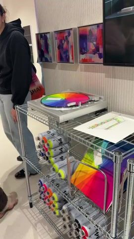 The video captures an indoor scene, likely an art exhibition or store, featuring colorful displays. In the foreground, a silver turntable with a rainbow-colored vinyl record is spinning on the top shelf of a metal shelving unit. Below, the shelves are filled with numerous spray paint cans, predominantly in neon yellow, orange, pink, and purple hues, many labeled "MONTANA COLORS 94" and "MONTANA COLORS". A piece of paper with a drawing or sketch, and a rainbow-striped folder, are also visible on the shelves.

A person wearing a black hoodie and light-colored jeans stands in the mid-ground, their back mostly to the camera. They are carrying a black tote bag which has text including "09.11-09.17", "2023", and "SHANGHAI". Behind the turntable, three framed abstract artworks with vibrant, varied colors hang on a white, textured wall. A portion of a TV screen displaying an image is visible on the far right. The overall setting appears to be a well-lit, contemporary space showcasing art-related items.
