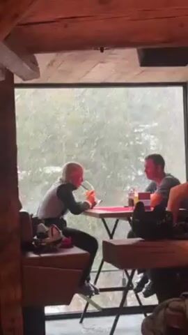 The video shows a woman with blonde hair, dressed in ski clothes, seated at a table in a rustic, wooden-framed building, drinking from a tall, stemmed glass with a straw. Seated at the same table is a man with short dark hair wearing a dark t-shirt. They appear to be having a conversation.  Outside the large picture window behind them, a snowstorm is occurring, obscuring the trees in the background. The table is covered with a red tablecloth. Ski gear is visible near the seating area.