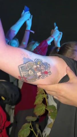 The video shows a person with a tattoo on their arm. The tattoo features cartoon characters. The person is at an outdoor event, likely a concert, with other people in the background. There is a large screen displaying animated characters. The event appears to be at night with stage lighting. The person holding the arm with the tattoo is smiling. The video is shaky and appears to be handheld.