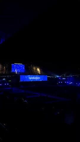 The video shows a large, dark venue, possibly a stadium or convention center, with a stage area illuminated by bright blue screens and spotlights. Two large screens are visible. The screen on the left displays a starry, cosmic background with some text. The screen on the right displays a similar starry background with the text "Agropecarios" and later "Forjada en valores," which translates to "Forged in values." There are bright bursts of light, possibly fireworks or confetti, erupting behind the stage area. The audience is dimly visible in the foreground, seated in rows. The overall impression is of a large-scale event, possibly an awards ceremony or conference, with a focus on a technological or agricultural theme.