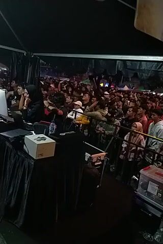 Here is a description of the video:

The video is shot from the side of a stage, looking out at a dense crowd of people. The angle is slightly tilted. The stage is covered in black fabric, and on it, a person is bent over, possibly interacting with equipment. The crowd is packed tightly together, with many individuals wearing hats or hoods. There is a tent-like structure overhead, providing cover for both the stage and the audience. Lighting is dim, contributing to an atmosphere of energy and excitement.