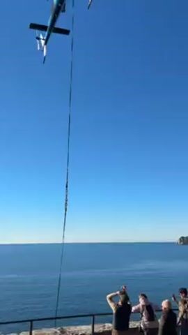 The video shows a turquoise and white helicopter hovering in the sky, positioned above a cliff overlooking the sea. A long cable hangs from the helicopter. Below, a group of people stand on the cliff, watching and taking pictures. The sea stretches out to the horizon, and a small island or headland is visible in the distance. The sky is clear and blue. The helicopter’s rotors are actively turning, indicating it is in operation. The overall scene suggests a planned event involving the helicopter and the group on the cliff.