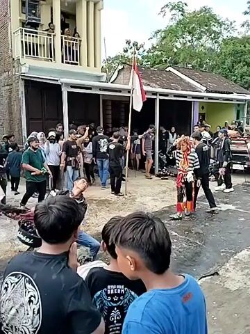 A group of people, some in traditional costumes, are gathered in an outdoor area. A person in a black and white striped costume with a colorful headdress is performing a dance. Other performers are seen with drums and musical instruments. There are spectators watching the performance. The environment appears to be a street or courtyard with buildings and trees in the background. The scene seems to be part of a cultural event or festival.