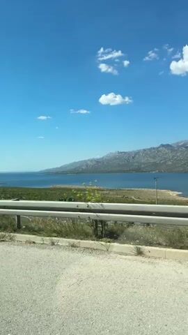 The video shows a view from a moving vehicle looking out at a body of water and mountains. In the foreground, there is a road and a guardrail. The water is blue and calm, and the mountains in the background are rocky and covered with sparse vegetation. The sky is a clear blue with a few scattered white clouds. The sun is shining, indicating it is daytime.