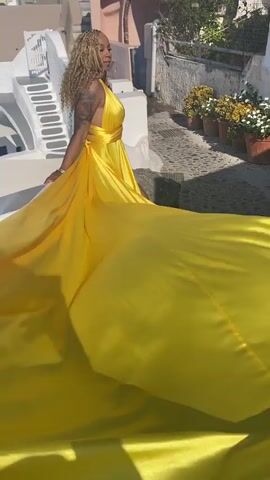 A woman with blonde curly hair, tattoos on her arms, and wearing a bright yellow halter dress with a long train stands on a white ledge overlooking a cobblestone pathway lined with potted flowers. The background features white and pink buildings with blue accents, characteristic of Mediterranean architecture. The woman is posed with her left hand on the ledge, looking to her left. The dress's train is spread out behind her, flowing across the white ledge and onto the pathway. The scene is bathed in sunlight, casting shadows.