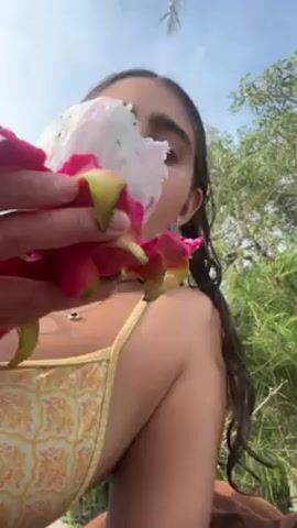 A woman in a yellow and orange floral print bathing suit holds up a flower to her face, then makes a kissy face at the camera. She has long brown hair, is wearing small silver earrings, and a necklace with a silver charm.  The sky is blue and there is green foliage in the background.