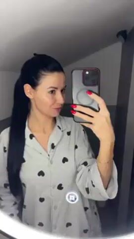 A woman with long, black hair tied into a ponytail is standing in front of a circular, illuminated mirror in what appears to be a bathroom or dressing area. She is wearing a gray pajama top with black heart patterns. She is holding a smartphone with a transparent case and red painted nails, taking a selfie. The background includes gray walls and a ceiling-mounted spotlight.