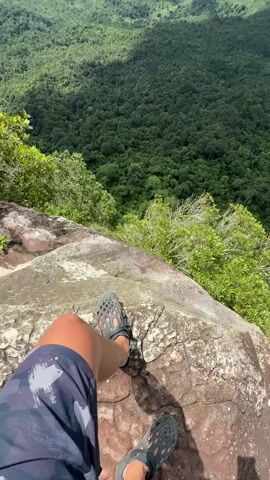 The video begins with a close-up shot of a person's legs and feet from a first-person perspective, sitting on a large rock formation. The person is wearing camouflage shorts and grey sandals. The camera then pans up to reveal a vast, lush green forest below, stretching out towards a hazy landscape of rolling hills and mountains in the distance. The sky above is partly cloudy with patches of blue. The viewpoint is from a high vantage point, likely a cliff or mountain peak, offering a breathtaking panoramic view of the natural scenery.