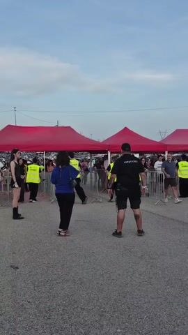 A security guard stands facing a crowd of people waiting behind metal barriers under red tents. People in yellow vests appear to be working. The ground is asphalt. In the background, there are more tents, a stage structure with lights, and a power line tower. The sky is cloudy. Several people are walking through the area.