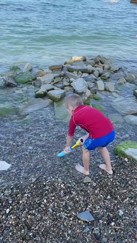A young boy wearing a red rash guard and blue swim trunks stands on a rocky shore near the ocean. He is holding a blue and yellow toy water gun, aiming it into the water. The boy is barefoot. The water is choppy with some waves breaking on the shore. The background shows more rocks, the ocean, and a distant cityscape with cranes and buildings.