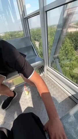 The video shows a view from inside a Ferris wheel cabin. In the foreground, a person's arm is visible, holding a phone to take a selfie. To the left, a plastic cup with a straw and what appears to be a drink is on the floor. To the right, a woman in a black top is seated, looking at the camera and smiling. She has a tattoo on her left arm and is wearing a bracelet. The cabin has metal flooring and gray seats. Through the large glass windows, a landscape of trees, buildings, a sports field, and the structure of the Ferris wheel itself can be seen under a blue sky with clouds. The camera pans slightly, showing different parts of the cabin and the outside view as the Ferris wheel moves.