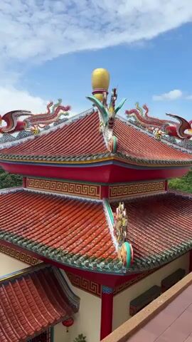 A close-up aerial shot pans across the ornate, traditional Chinese-style rooftops of a temple or shrine. The rooftops are covered in green ceramic tiles and adorned with colorful decorative elements, including red dragons and phoenixes. The sky above is blue with scattered white clouds. In the background, lush green trees are visible, suggesting the temple is situated in a natural setting. The camera movement is smooth and steady, offering a detailed view of the architectural features and the overall atmosphere of the location.