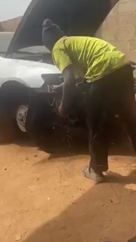 A man is working on a car with its hood up. The car is silver and appears to be missing its front bumper. The man is wearing a light green shirt, dark pants, a dark beanie, and sandals. He is standing in front of the car, reaching into the engine compartment. The ground appears to be dirt. A wall is visible in the background.