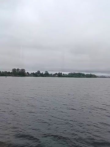 A cloudy day over a body of water. The water has small waves. In the distance, across the water, is a shoreline with trees and buildings. The shoreline appears to be a village or small town. There is no visible movement or action in the video.