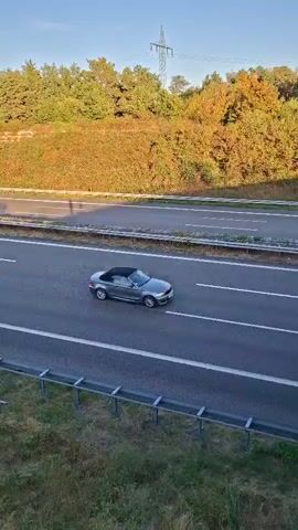 The video shows a highway scene with multiple vehicles. In the foreground, a red semi-trailer truck is visible, followed by a white car. Further down the highway, a light green and white truck with the logo "LUMPRECHT" is driving. Behind it, another white truck is seen. Another semi-trailer truck, this one blue, is in the right lane, appearing to be a dump truck. The background consists of a wooded hill with autumn foliage and a clear blue sky. A power line tower is also visible against the sky. The overall scene depicts a sunny day on a busy highway.