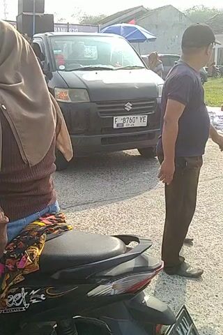 A dark-colored Suzuki Carry van is parked on a paved surface. A man wearing a dark blue t-shirt with white lettering that says "SATPAM" is standing next to the van, with his right hand on the hood. The van has a license plate that reads "F 8760 YE". In the foreground, a person's arm and hand are visible, resting on what appears to be a motorcycle seat. A person wearing a brown headscarf and a patterned garment is also visible in the foreground. To the left of the van, several large speakers are stacked. The background shows a building and some trees. The scene appears to be outdoors, with daylight.