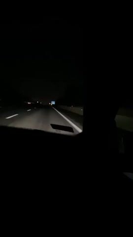 The video shows a nighttime view from inside a car driving on a highway. The headlights illuminate the road ahead, with white lines marking the lanes. Distant lights of other vehicles and signs are visible in the darkness. The right side of the frame is largely obscured by the dark silhouette of a person's head and shoulder. The overall scene conveys a sense of travel in the dark.