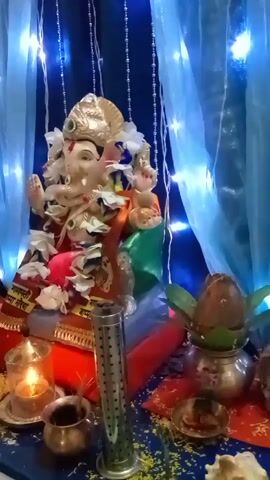 This is a short video of a decorated idol of Lord Ganesha. The idol is adorned with flowers and is seated on a colorful platform. In front of the idol, there is a lit diya (oil lamp), a small metal pot, and a decorative metal stand. To the right, there is a silver pot with a coconut, and some marigold petals are scattered around the altar. The background is decorated with blue lights and light blue curtains. The overall setting appears to be an altar or a shrine prepared for a religious occasion.