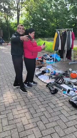 A man and a child are standing on a paved area and holding phones. In front of them is a collection of items laid out on the ground, including clothing, shoes, electronics, and household items. A clothing rack with clothes is behind them. The environment appears to be outdoors, possibly a flea market or garage sale, with trees and a road in the background.