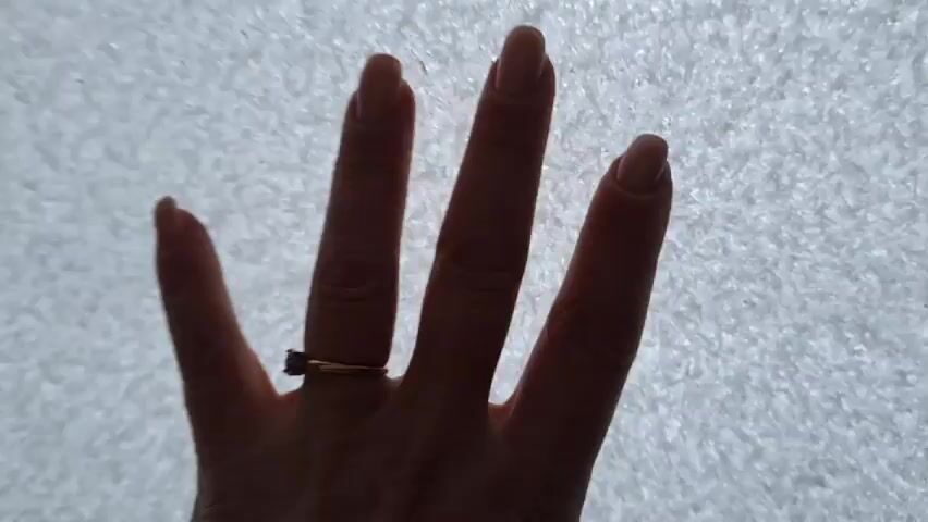 A hand with a ring brushes snow off a window. The fingers are visible, and there is a gold ring on the ring finger. The hand moves across the window, clearing a path through the snow. The snow appears to be fresh and light.