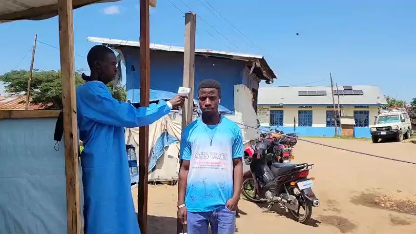 In Tchere, Democratic Republic of the Congo, a bright daylight scene unfolds on a dusty ground. Two men are captured in the frame, engaged in what appears to be a health screening.

On the left, a man dressed in a light blue, full-length garment, possibly a medical scrub or traditional attire, holds an infrared thermometer gun. He is standing under a simple wooden structure, which seems to be part of a makeshift stall or shelter. His focus is directed towards the second man.

The second man stands in the center of the image, facing the camera with a neutral expression. He is wearing a light blue t-shirt with "OASIS WATER" prominently displayed on the front, along with smaller, less legible text underneath. He is also wearing blue jeans. Around his neck, he has earbuds or a small headset.

Behind the man being screened, a black and red motorcycle is parked on the dirt ground. In the background, under a clear blue sky, there are buildings. One larger building, painted light yellow and blue, has solar panels visible on its roof. To the right of this building, a white utility truck is parked. The overall setting suggests a semi-rural or developing community, with basic infrastructure and the presence of both traditional elements (wooden structures) and modern technologies (solar panels, motorcycle). The activity of temperature taking indicates a health-related measure, possibly a COVID-19 screening or general health check.