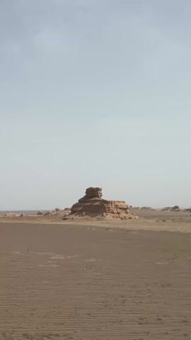 The video displays a vast, desolate desert landscape under a hazy, bright sky. Unique, erosion-formed rock structures, known as yardangs, are scattered across the sandy terrain, appearing as isolated mounds and ridges in the distance. The ground in the foreground shows distinct parallel lines, possibly tire tracks or wind-formed ripples. The camera slowly pans from right to left, revealing more of the expansive desert and its sparse, rugged features. The scene suggests a dry, arid environment, likely in the daytime given the soft, widespread light. This landscape is characteristic of the Gobi Desert near Dunhuang City, China. No people or text are visible.