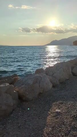 The video shows a view of the sea from a rocky shore during sunset. The sun is visible in the upper right quadrant of the frame, casting a reflection on the water. The sky is a light blue with some scattered clouds. In the background, there are mountains. The foreground is made up of rocks and gravel. There are no people or animals visible in the video. The water appears to be calm with small waves.