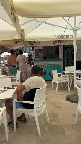 The video opens with a close-up of a white outdoor table set at what appears to be a cafe or restaurant. A man is seated in a white chair facing away from the camera. He is wearing a white t-shirt and blue shorts, with a black backpack on the back of his chair. Sunglasses rest on the table in front of him, next to a menu. The camera pans to the right, revealing more white tables and chairs under large, cream-colored umbrellas. In the background, a counter with a menu board and an ice cream freezer is visible. Several people are standing and interacting near the counter. The umbrellas are branded with "Carlsberg" and "TUBORG" logos, suggesting a beverage sponsor. As the camera continues to pan right and slightly upward, the scene expands to show a beach area with lounge chairs and umbrellas. People are visible relaxing on the beach and in the water. The sea stretches out to the horizon, with a lush green mountainside in the distance. The overall impression is of a sunny day at a seaside establishment.