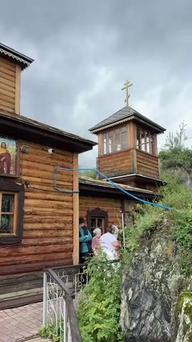 A video shows a wooden church with a bell tower. Three people are standing outside the church. The church has several windows, religious icons, and a cross on top of the bell tower. The sky is cloudy.