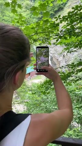 A person is holding a smartphone and filming a waterfall. The phone is in portrait mode. The person is using their fingers to interact with the phone screen. The background shows a waterfall cascading down a rocky cliff face into a pool of water. There is also a small wooden building with a green roof and stairs near the waterfall. Lush green foliage surrounds the scene. The time stamp on the phone screen reads 00:00:01.