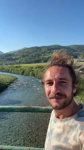 A man with a beard, mustache, and a bun hairstyle stands on a bridge with a green railing, smiling and looking at the camera. Behind him is a river, green grass, hills, and a clear blue sky. He is wearing a light-colored tie-dye shirt. A woman in a white shirt comes into view and briefly stands next to the man. They kiss. The video then shows the other side of the bridge, with a road, trees, buildings, and hills in the background. The sun is shining brightly.