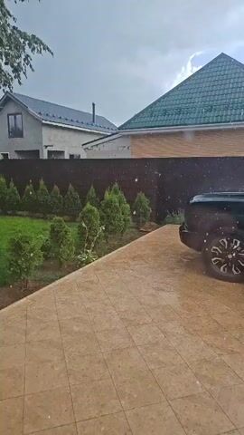 The video captures an outdoor scene, possibly a backyard or driveway, with a paved area in the foreground. A black SUV is parked to the right of the frame. Beyond the paved area, a neatly manicured lawn is visible, bordered by a row of small evergreen trees. Further back, a dark fence separates the lawn from neighboring properties. Two houses are partially visible behind the fence, one with a grey facade and dark roof, the other with a brick-colored lower section and a green, sloped roof. The sky appears overcast, and what appear to be large raindrops are visible falling throughout the scene, suggesting it is raining.