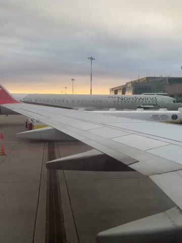 This media file captures a view from an airplane window, likely at an airport in Stolbishche, Russia. Two Nordwind Airlines airplanes are parked side-by-side on the tarmac. The aircraft are predominantly white with red tail fins featuring the Nordwind Airlines logo, a stylized 'N'. Visible text on the side of one aircraft includes "NORDWIND Airlines" and what appears to be a registration number starting with "RA-".

The scene depicts airport operations. Various ground vehicles, including a baggage tug and a small loader, are positioned near the planes, suggesting that baggage handling or preparation for flight is underway. Orange traffic cones are also visible on the tarmac. The sky is overcast with a soft glow of sunrise or sunset visible on the horizon, indicating it is either early morning or late evening. The overall mood is calm, typical of pre-flight or post-flight operations. The view is dominated by the wing of the airplane the recording is from, with the tail section and fuselage of the adjacent aircraft clearly visible.